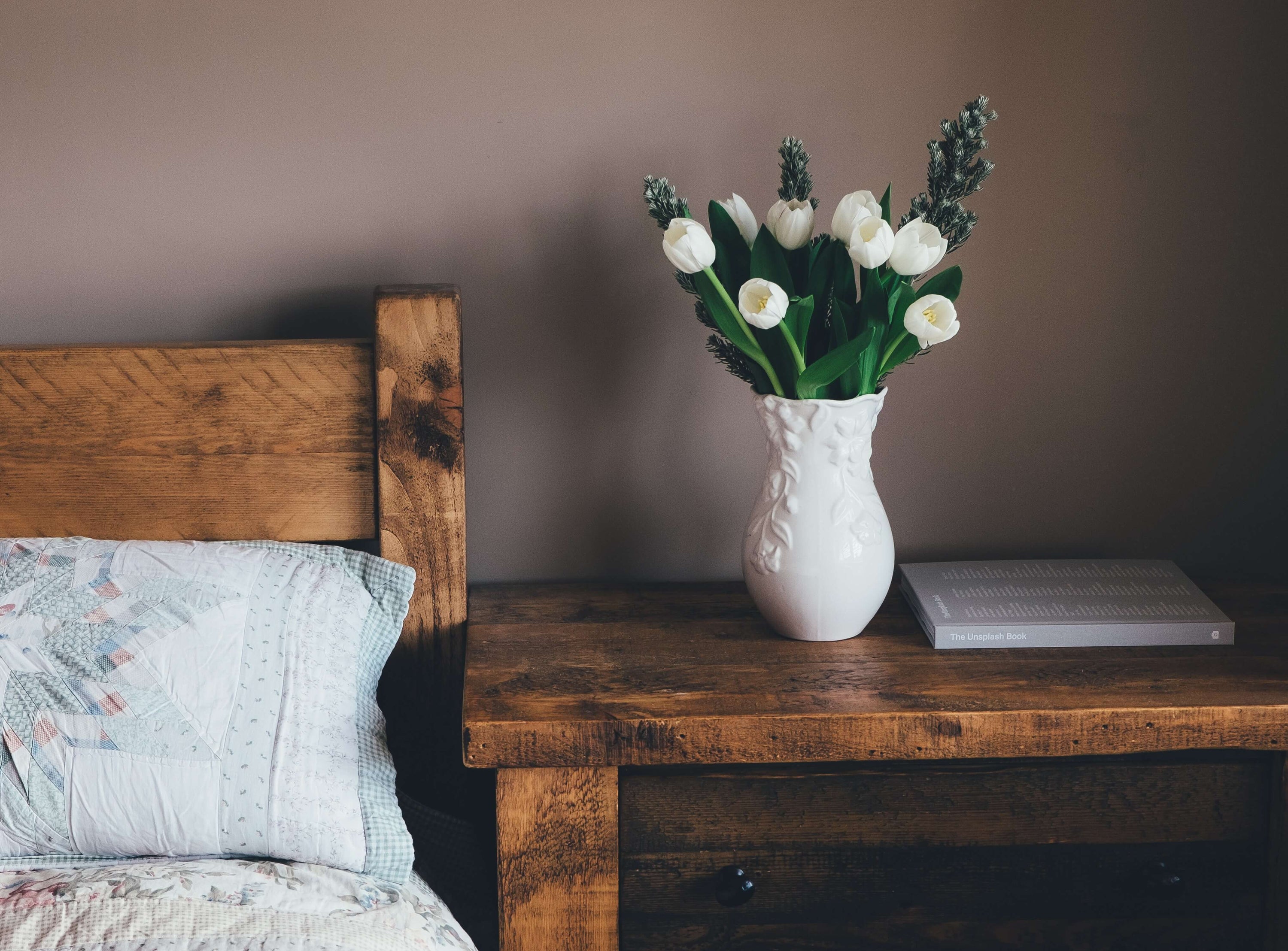 Vase of white flowers on a rustic wooden table next to a comfortable bed
