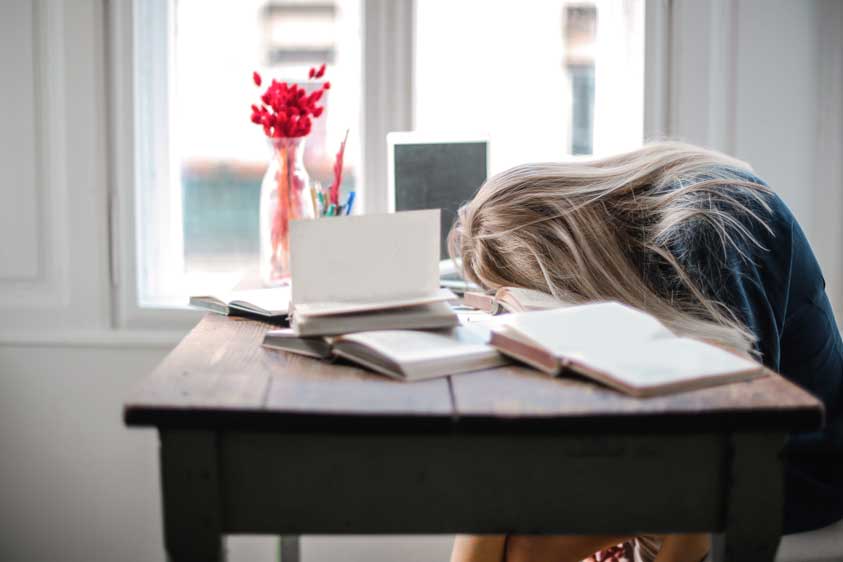  A woman sits asleep in her chair with her head on her desk, surrounded by books.